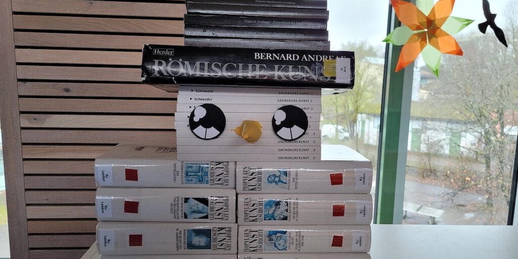 Ein Stapel Bücher, die so angeordnet sind, dass sie einem Gesicht ähneln, mit Comic-Augen und einem Schnabel. Das Arrangement steht auf einem Tisch in der Nähe eines Fensters mit einer bunten Sternendekoration. Die Bücher haben schwarze, weiße und graue Einbände.