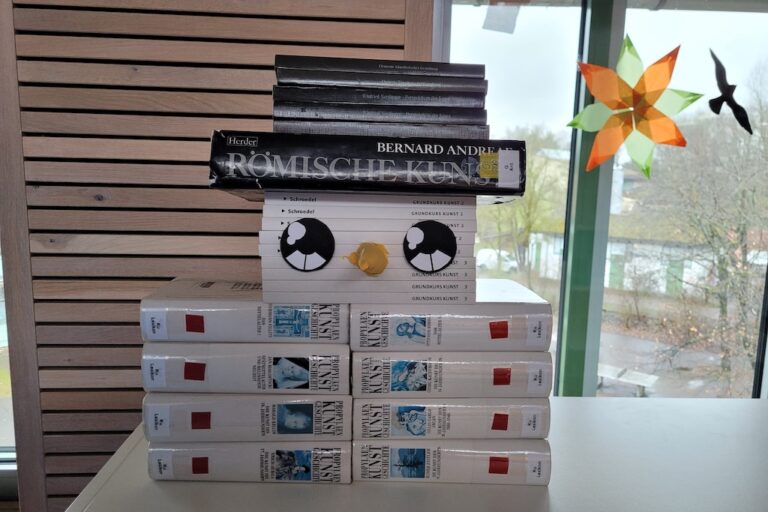 Ein Stapel Bücher, die so angeordnet sind, dass sie einem Gesicht ähneln, mit Comic-Augen und einem Schnabel. Das Arrangement steht auf einem Tisch in der Nähe eines Fensters mit einer bunten Sternendekoration. Die Bücher haben schwarze, weiße und graue Einbände.