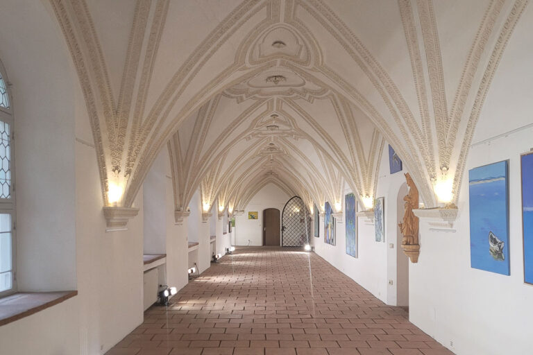 Ein langer, gewölbter Flur mit kunstvollen Gewölbedecken, Kunstwerken an weißen Wänden und sanftem, warmem Licht. Links befinden sich Fenster und rechts Statuen.