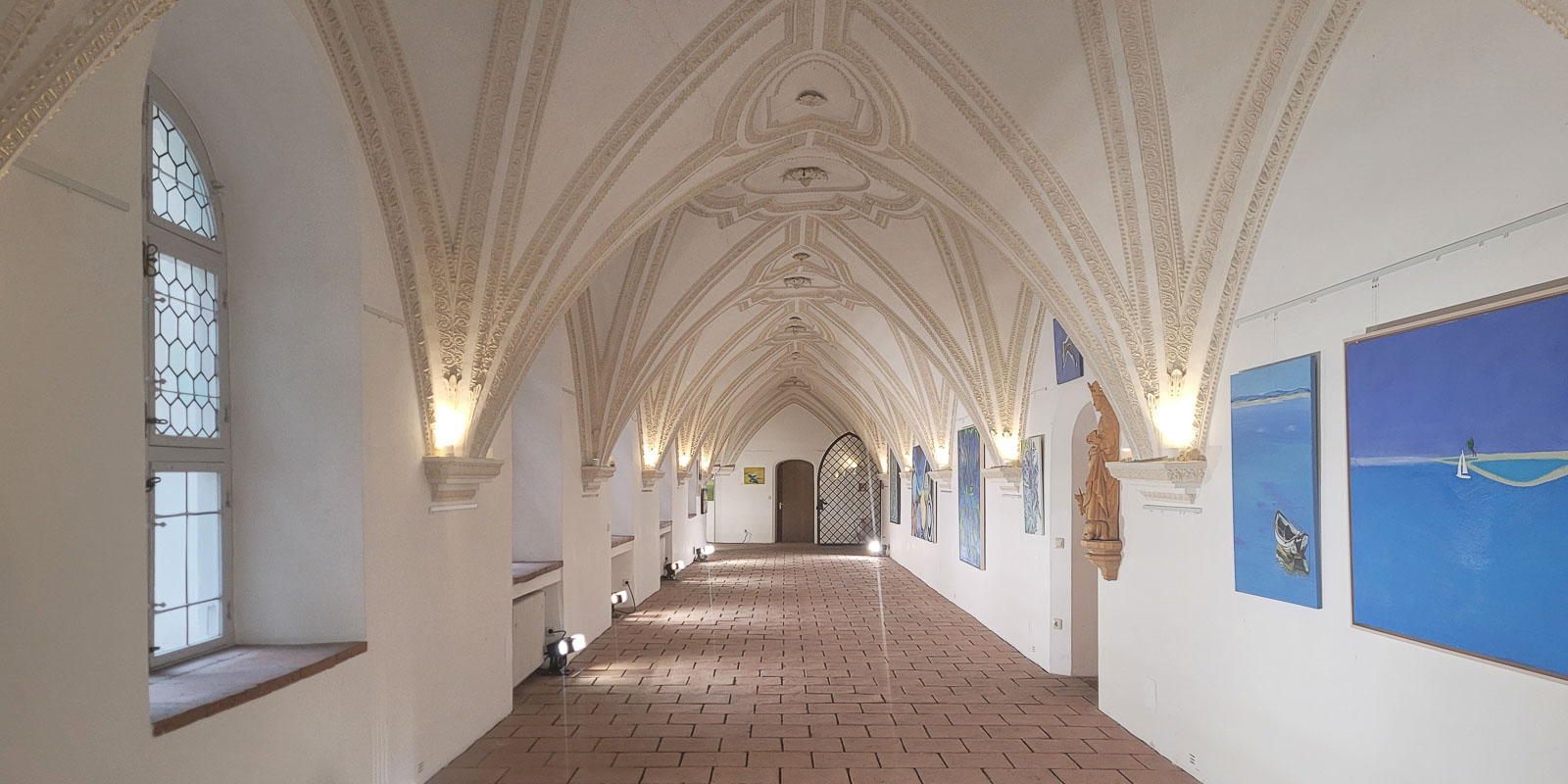 20250312_092914-4 Ein langer, gewölbter Flur mit kunstvollen Gewölbedecken, Kunstwerken an weißen Wänden und sanftem, warmem Licht. Links befinden sich Fenster und rechts Statuen.