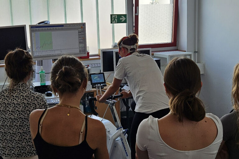 Vier Personen beobachten, wie eine Person auf einem stationären Fahrrad in einem Labor einen Fitnesstest absolviert, während im Hintergrund medizinische Geräte und Monitore die Daten anzeigen.