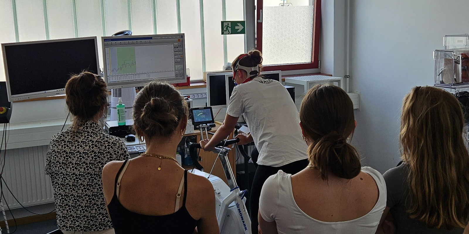 20250623_130336-2 Vier Personen beobachten, wie eine Person auf einem stationären Fahrrad in einem Labor einen Fitnesstest absolviert, während im Hintergrund medizinische Geräte und Monitore die Daten anzeigen.