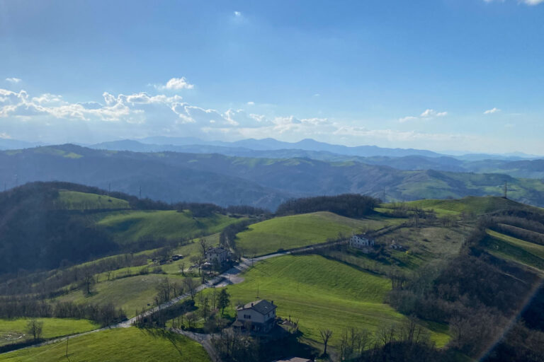 Eine malerische Aussicht auf sanfte grüne Hügel und verstreute Häuser unter einem klaren blauen Himmel, mit Bergen in der Ferne, die im Hintergrund verschwinden, und Licht- und Schattenflecken, die sich über die Landschaft erstrecken.