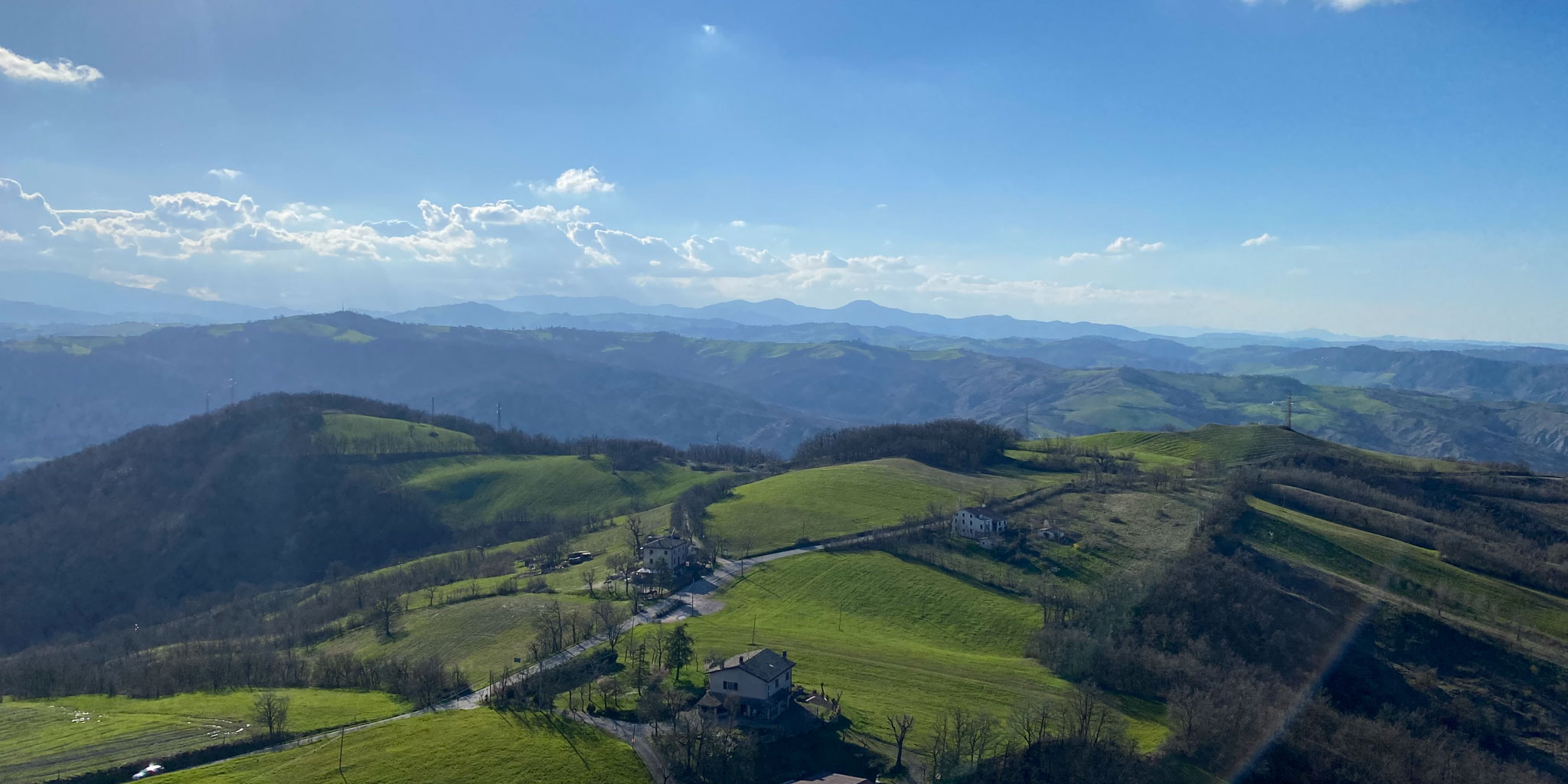 Aussicht vom Castello di Rossena Eine malerische Aussicht auf sanfte grüne Hügel und verstreute Häuser unter einem klaren blauen Himmel, mit Bergen in der Ferne, die im Hintergrund verschwinden, und Licht- und Schattenflecken, die sich über die Landschaft erstrecken.