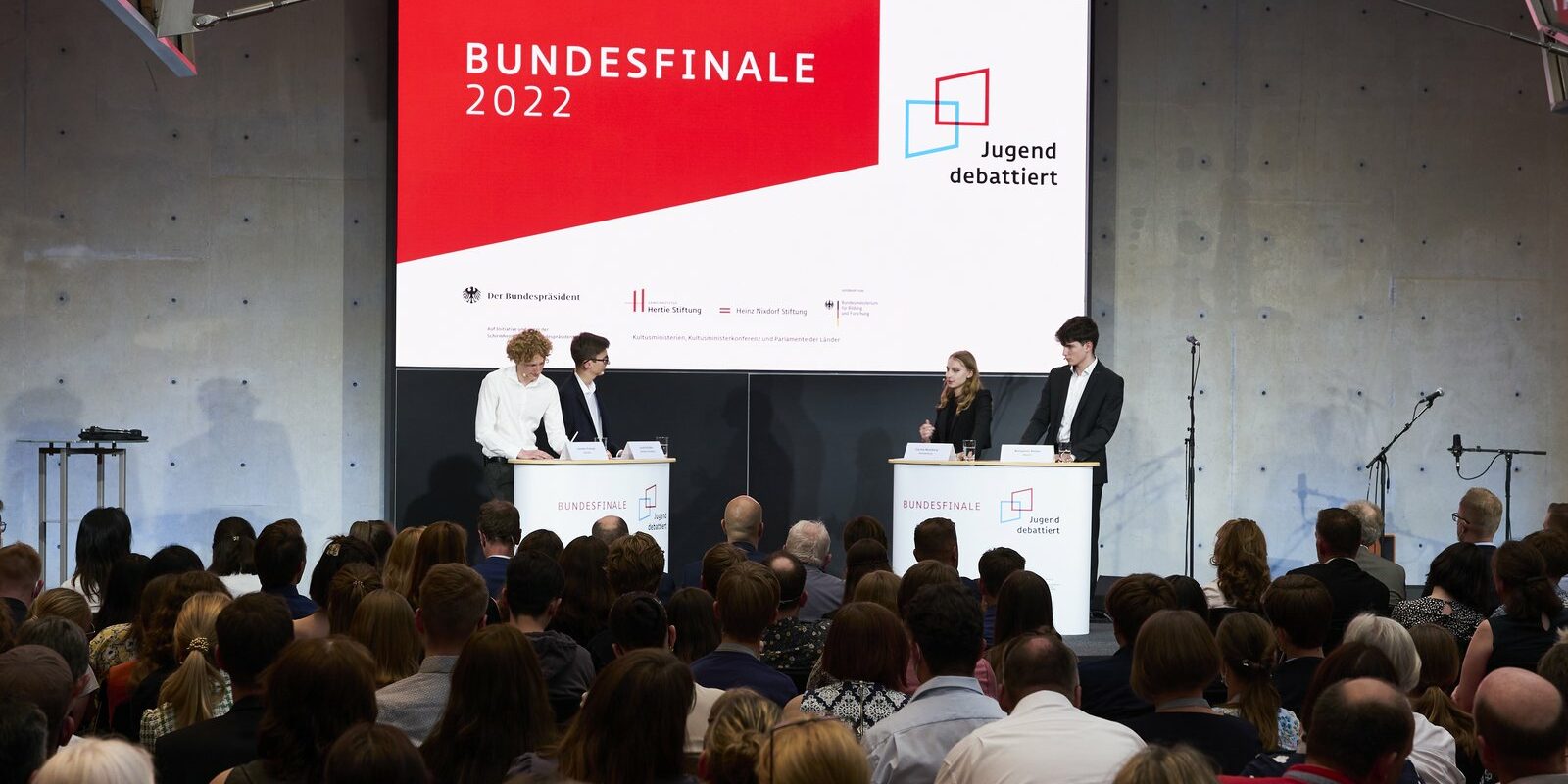 BFJD_2022_18062022_2136 Auf der Bühne findet ein Debattierwettbewerb mit vier Teilnehmern an Podien statt. Auf einer großen Leinwand laufen die Titel „Bundesfinale 2022“ und „Jugend debattiert“. Das Publikum sitzt und verfolgt aufmerksam das Geschehen.