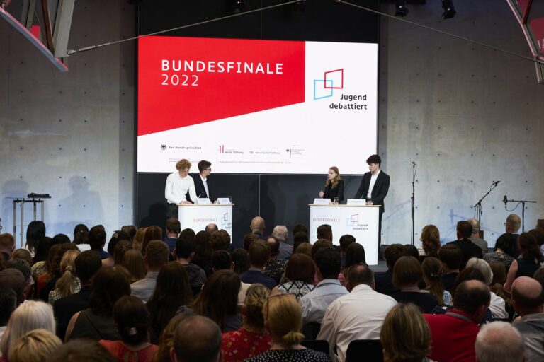 Auf der Bühne findet ein Debattierwettbewerb mit vier Teilnehmern an Podien statt. Auf einer großen Leinwand laufen die Titel „Bundesfinale 2022“ und „Jugend debattiert“. Das Publikum sitzt und verfolgt aufmerksam das Geschehen.