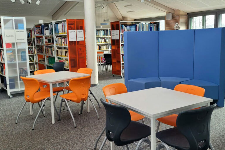 Eine moderne Bibliothek mit Bücherregalen, zwei Tischen mit orangefarbenen Stühlen, einem Tisch mit schwarzen Stühlen und einer geschwungenen blauen Sitzecke. Der Raum ist dank großer Fenster gut beleuchtet.