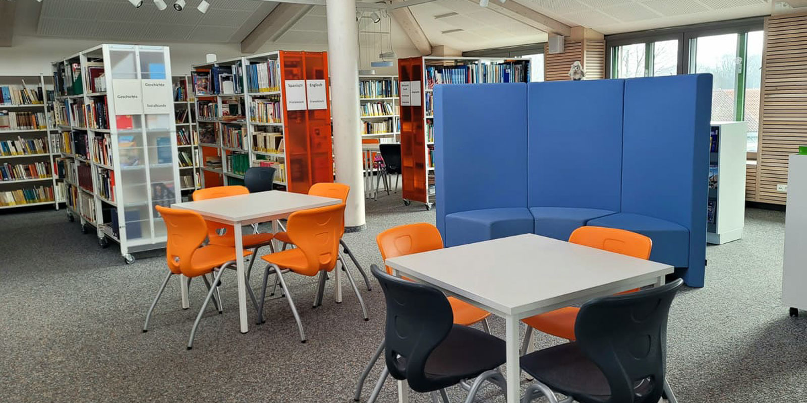 Bibliothek-1-2 Eine moderne Bibliothek mit Bücherregalen, zwei Tischen mit orangefarbenen Stühlen, einem Tisch mit schwarzen Stühlen und einer geschwungenen blauen Sitzecke. Der Raum ist dank großer Fenster gut beleuchtet.