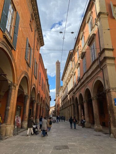 Bologna-6 Menschen gehen unter einem teilweise bewölkten Himmel eine Kopfsteinpflasterstraße entlang, die von historischen orange- und beigefarbenen Gebäuden mit Bögen und Säulen gesäumt ist. Im Hintergrund erhebt sich ein hoher mittelalterlicher Turm.