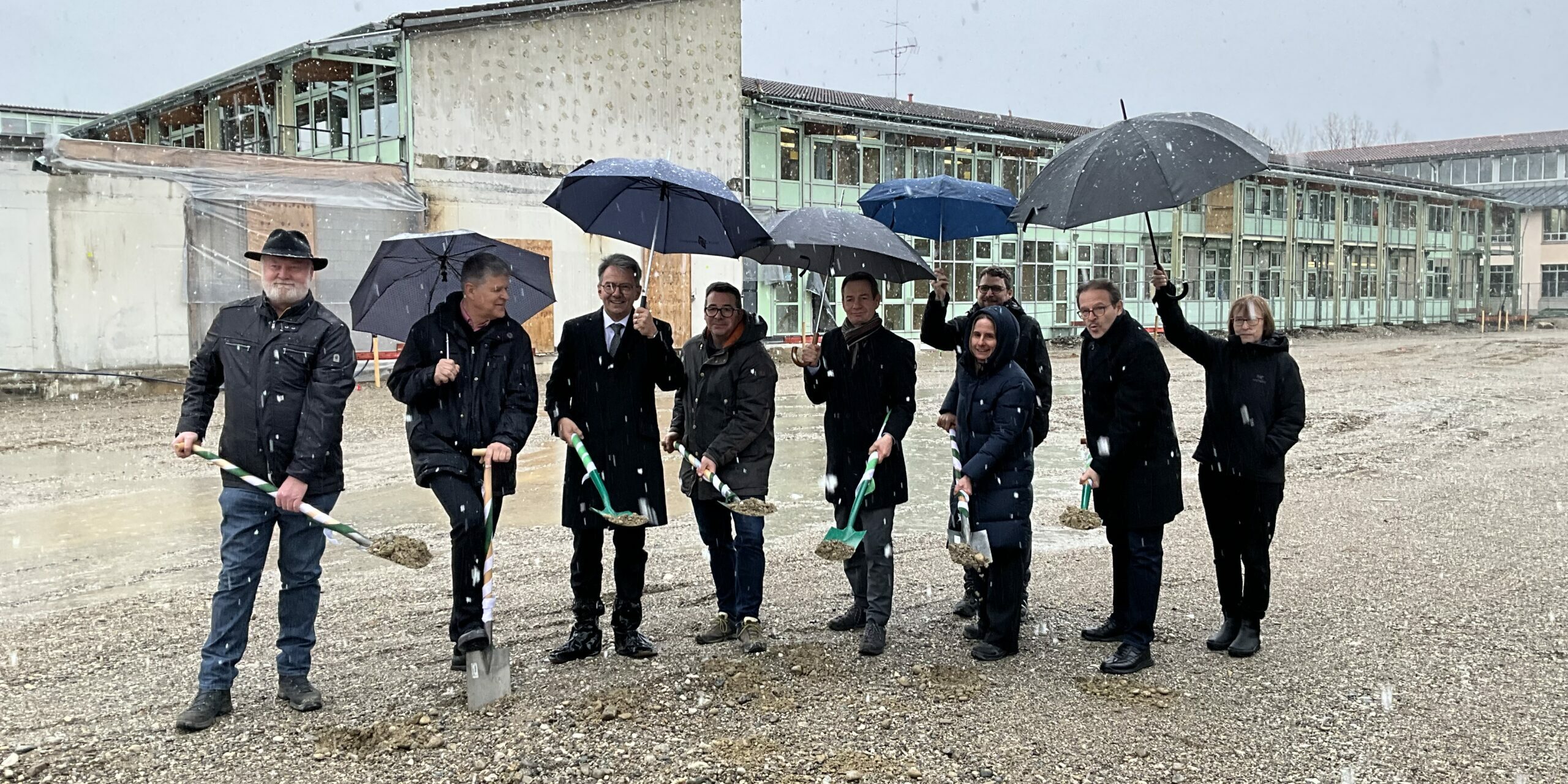 D5EFBFF7-E439-4639-A22E-B4FADB424D51 Neun Menschen in Winterkleidung stehen an einem verschneiten Tag im Freien und halten Schaufeln und Regenschirme in den Händen. Sie scheinen an einer Grundsteinlegungszeremonie vor einem alten Gebäude teilzunehmen.