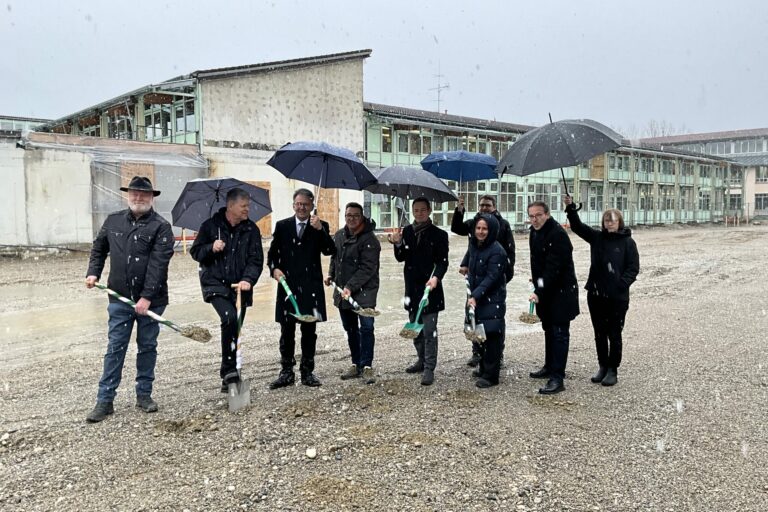 Neun Menschen in Winterkleidung stehen an einem verschneiten Tag im Freien und halten Schaufeln und Regenschirme in den Händen. Sie scheinen an einer Grundsteinlegungszeremonie vor einem alten Gebäude teilzunehmen.