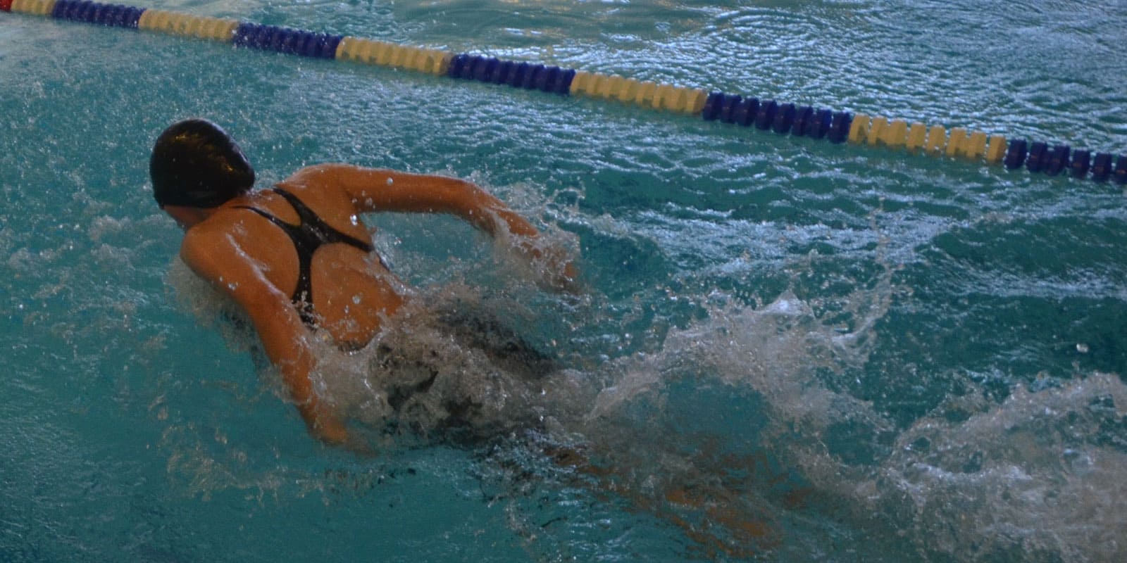 DSC_2376-4 Ein Schwimmer, der eine schwarze Badekappe und einen Badeanzug trägt, schwimmt Freistil in einem Schwimmbecken und verursacht Spritzer, während er sich neben einer Bahntrennung bewegt.