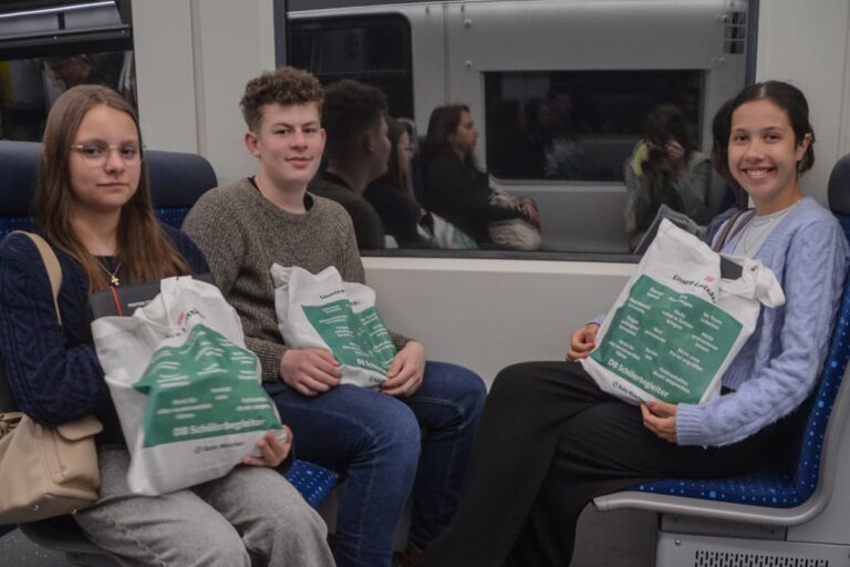 Drei junge Menschen sitzen mit ähnlichen weiß-grünen Tragetaschen in einem Zug. Die beiden linken haben einen neutralen Gesichtsausdruck, die rechte Person lächelt. Im Hintergrund sitzen weitere Passagiere.