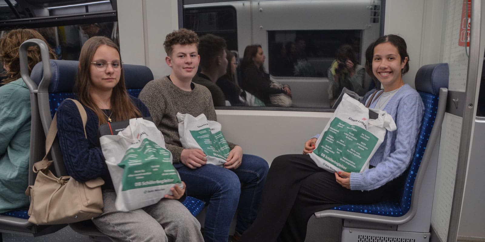 DSC_3392-2 Drei junge Menschen sitzen mit ähnlichen weiß-grünen Tragetaschen in einem Zug. Die beiden linken haben einen neutralen Gesichtsausdruck, die rechte Person lächelt. Im Hintergrund sitzen weitere Passagiere.