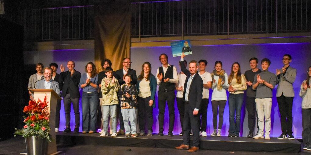 Forscherschule_Preisverleihung Eine Gruppe von Menschen steht auf einer Bühne, einige halten Urkunden in den Händen. Auf zwei großen Bannern an beiden Seiten steht „jugend forscht“. Vor der Bühne steht ein Podium mit roten Blumen. Das Publikum scheint einer Präsentation oder Preisverleihung zuzuschauen.