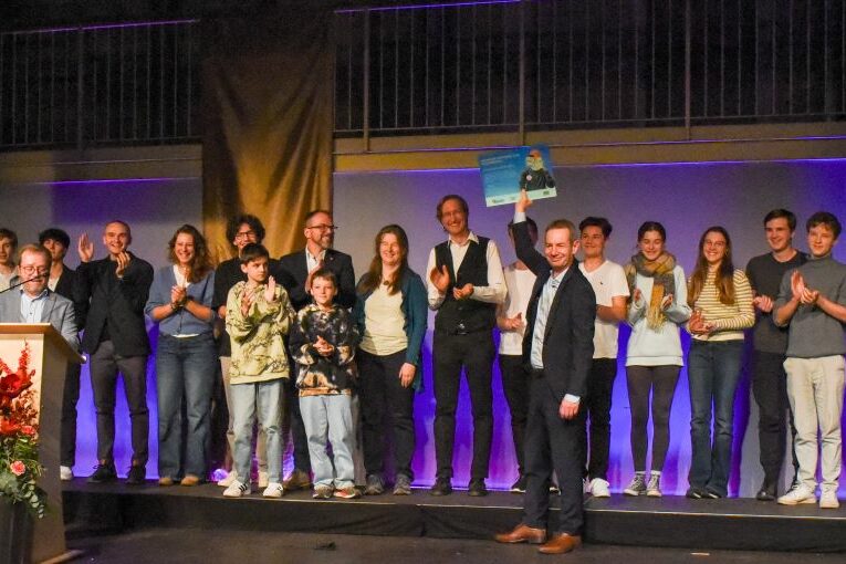 Eine Gruppe von Menschen steht auf einer Bühne, einige halten Urkunden in den Händen. Auf zwei großen Bannern an beiden Seiten steht „jugend forscht“. Vor der Bühne steht ein Podium mit roten Blumen. Das Publikum scheint einer Präsentation oder Preisverleihung zuzuschauen.