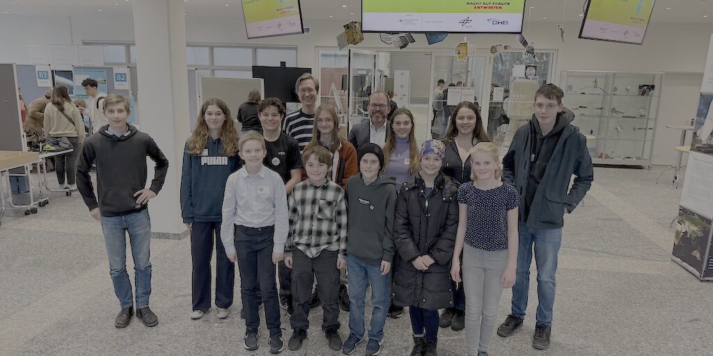 Eine Gruppe von vierzehn Personen, darunter Erwachsene und Kinder, steht in einem großen, modernen Raum mit Displays und Bildschirmen im Hintergrund. Sie alle blicken in die Kamera und lächeln.