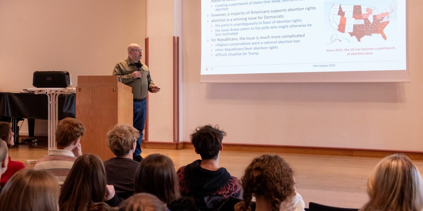 Eine Person hält vor einem Publikum einen Vortrag, wobei auf einem Projektor eine Folie über Abtreibungsgesetze angezeigt wird. Die Folie enthält Text und eine Karte der US-Bundesstaaten, auf der die Unterschiede in den Abtreibungsgesetzen hervorgehoben werden. Das Publikum sitzt und hört aufmerksam zu.