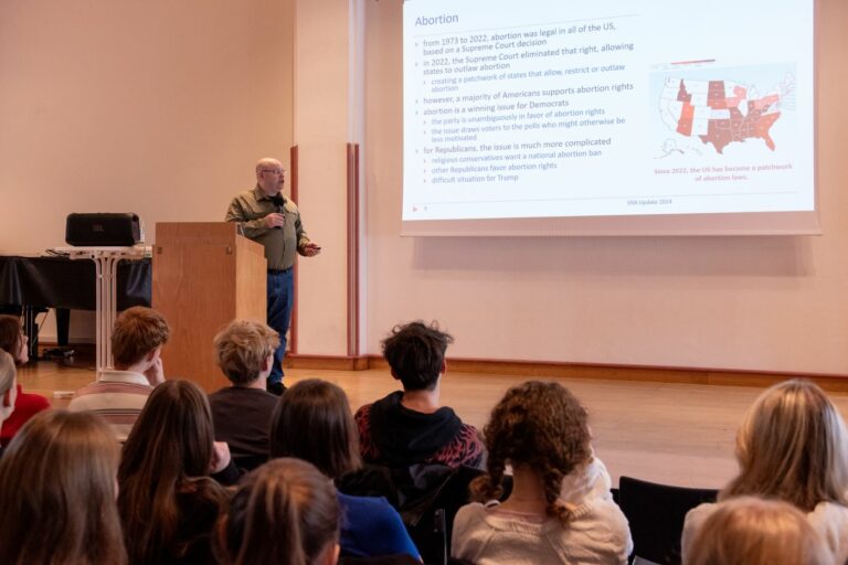 Eine Person hält vor einem Publikum einen Vortrag, wobei auf einem Projektor eine Folie über Abtreibungsgesetze angezeigt wird. Die Folie enthält Text und eine Karte der US-Bundesstaaten, auf der die Unterschiede in den Abtreibungsgesetzen hervorgehoben werden. Das Publikum sitzt und hört aufmerksam zu.