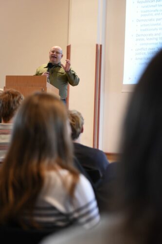 Ein Redner im grünen Hemd hält einen Vortrag vor Publikum. Auf der Projektionsfläche wird Text angezeigt. Der Fokus liegt auf dem Redner am Podium, im Vordergrund das Publikum.