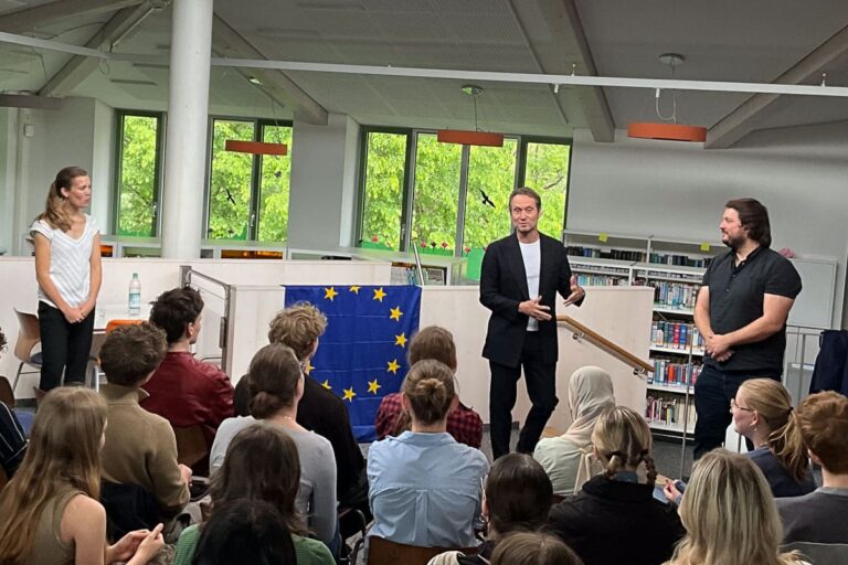 Ein Mann in einem dunklen Anzug spricht zu einem Publikum in einer Bibliothek, flankiert von einer Frau auf der linken und einem anderen Mann auf der rechten Seite. Im Hintergrund steht ein Tisch mit einer EU-Flagge. Die Zuhörer sitzen und hören aufmerksam zu.