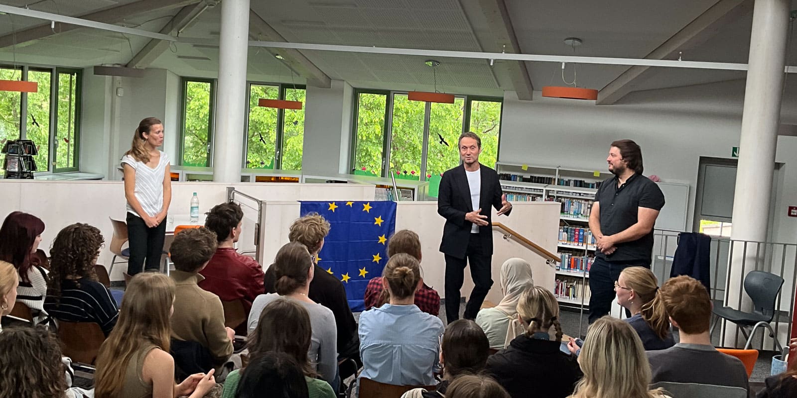 IMG_0673 Ein Mann in einem dunklen Anzug spricht zu einem Publikum in einer Bibliothek, flankiert von einer Frau auf der linken und einem anderen Mann auf der rechten Seite. Im Hintergrund steht ein Tisch mit einer EU-Flagge. Die Zuhörer sitzen und hören aufmerksam zu.