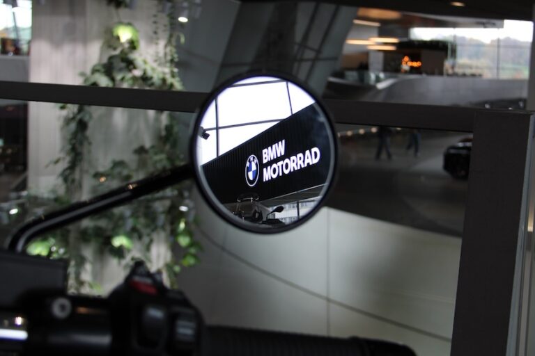 Im Rückspiegel eines Motorrads spiegelt sich das BMW Motorrad Logo. Im Vordergrund ist der Lenkergriff zu sehen, im Hintergrund sind das Innere eines Gebäudes und etwas Grün zu sehen.