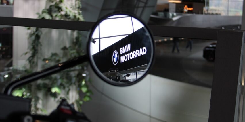 Im Rückspiegel eines Motorrads spiegelt sich das BMW Motorrad Logo. Im Vordergrund ist der Lenkergriff zu sehen, im Hintergrund sind das Innere eines Gebäudes und etwas Grün zu sehen.