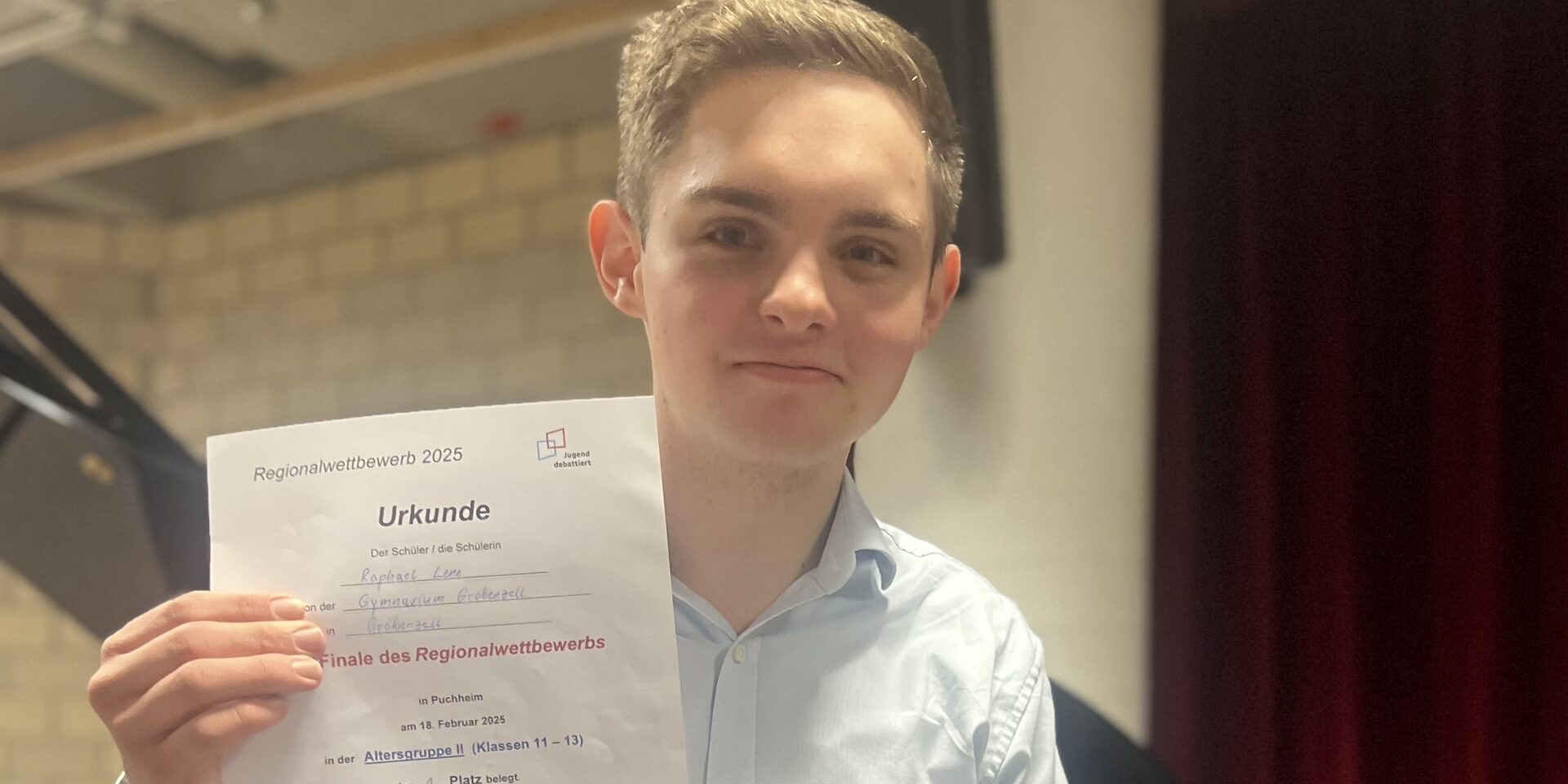 IMG_5848 4 Eine junge Person mit kurzen Haaren und hellblauem Hemd lächelt und hält eine Urkunde hoch. Im Hintergrund sind eine Backsteinmauer und Theatervorhänge zu sehen. Die Urkunde enthält einen deutschen Text, der sich auf einen Regionalwettbewerb im Jahr 2023 bezieht.