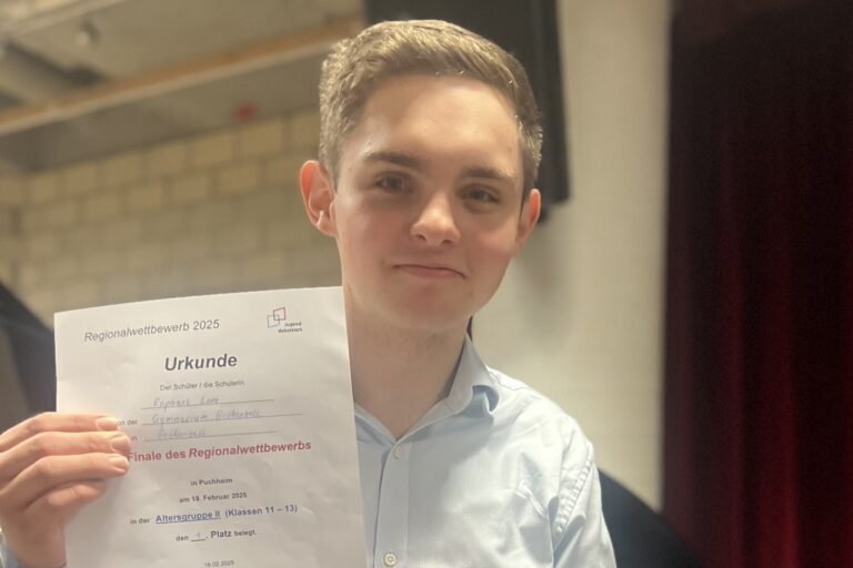 Eine junge Person mit kurzen Haaren und hellblauem Hemd lächelt und hält eine Urkunde hoch. Im Hintergrund sind eine Backsteinmauer und Theatervorhänge zu sehen. Die Urkunde enthält einen deutschen Text, der sich auf einen Regionalwettbewerb im Jahr 2023 bezieht.