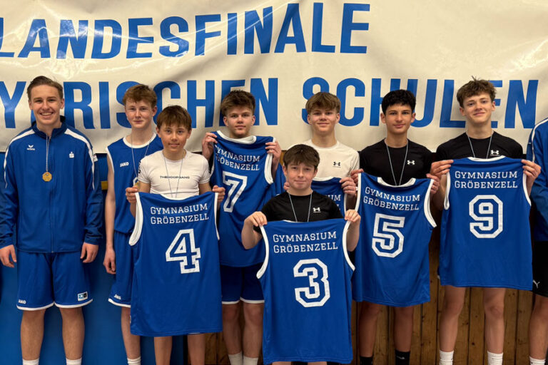Eine Gruppe von zehn Jungen in einheitlichen Sportuniformen steht vor einem Banner mit der Aufschrift „Landesfinale Bayerischen Schulen“. Sie halten Basketballtrikots mit der Aufschrift „Gymnasium Gröbenzell“, lächeln und tragen Medaillen.