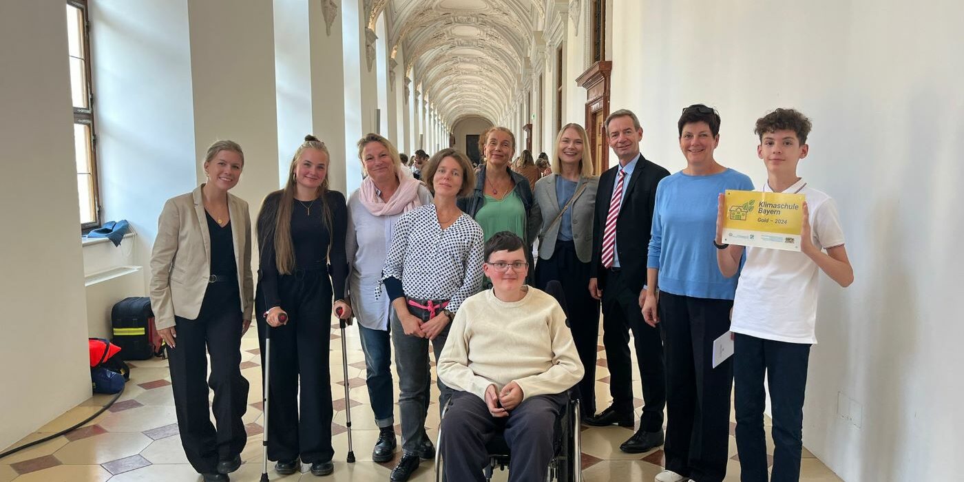 KlimaschuleGold3 Eine Gruppe von zwölf Personen, darunter eine Person im Rollstuhl und ein junger Mensch, der ein gelbes Schild hält, posiert gemeinsam in einem langen, eleganten Flur mit dekorativen Decken und großen Fenstern. Die meisten lächeln und sind leger gekleidet.