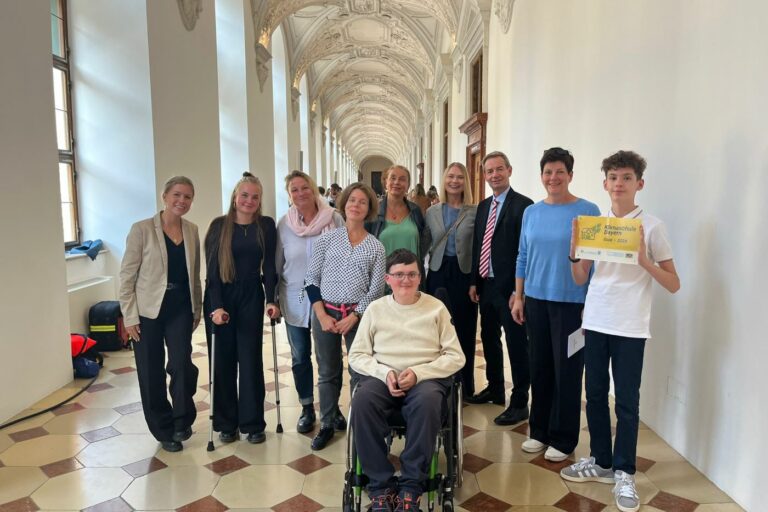 Eine Gruppe von zwölf Personen, darunter eine Person im Rollstuhl und ein junger Mensch, der ein gelbes Schild hält, posiert gemeinsam in einem langen, eleganten Flur mit dekorativen Decken und großen Fenstern. Die meisten lächeln und sind leger gekleidet.