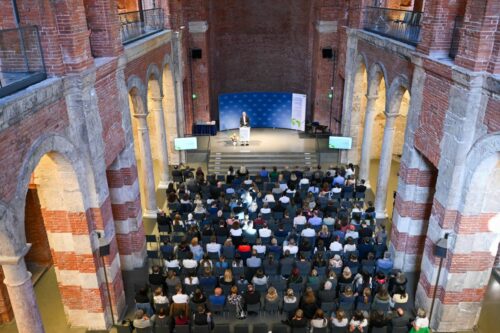 Ein Redner hält einen Vortrag in einem großen, historischen Backsteinsaal mit hohen Decken. Das Publikum sitzt vor einer Bühne, die auf beiden Seiten mit Projektionsbildschirmen ausgestattet ist.