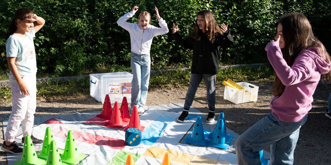 Popup Vier Kinder spielen ein Outdoor-Spiel auf einer großen, bunten Matte mit grünen, roten und orangefarbenen Kegeln. Sie wirken aufgeregt und stehen an verschiedenen Ecken, im Hintergrund ist ein Waldgebiet zu sehen.