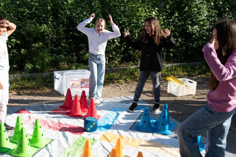 Vier Kinder spielen ein Outdoor-Spiel auf einer großen, bunten Matte mit grünen, roten und orangefarbenen Kegeln. Sie wirken aufgeregt und stehen an verschiedenen Ecken, im Hintergrund ist ein Waldgebiet zu sehen.
