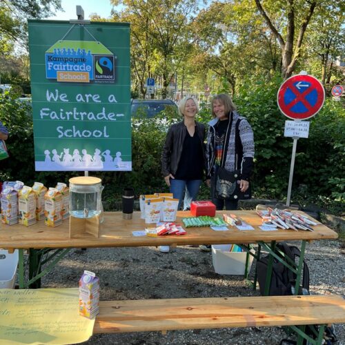 Zwei Personen stehen bei einer Veranstaltung im Freien hinter einem Tisch, der für eine Fairtrade-Schule wirbt. Auf dem Tisch sind verschiedene Fairtrade-Produkte wie Saftkartons und Snacks ausgestellt. Auf einem großen Schild im Hintergrund steht: „Wir sind eine Fairtrade-Schule.“ Dahinter sind Bäume zu sehen.