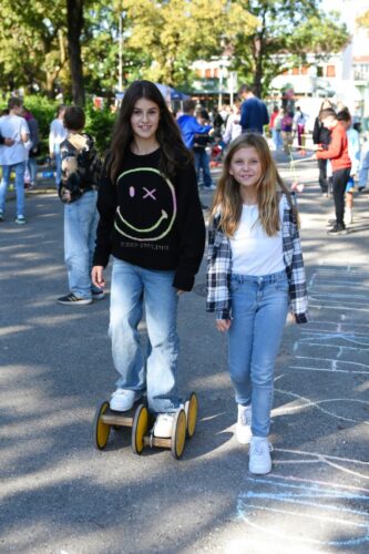 Zwei Mädchen genießen einen sonnigen Tag in einem Park. Eines fährt auf einem kleinen gelben Balance Board und trägt einen Pullover mit Smiley, während die andere in einem Flanellhemd neben ihr läuft. Im Hintergrund sind Menschen und Grün zu sehen.