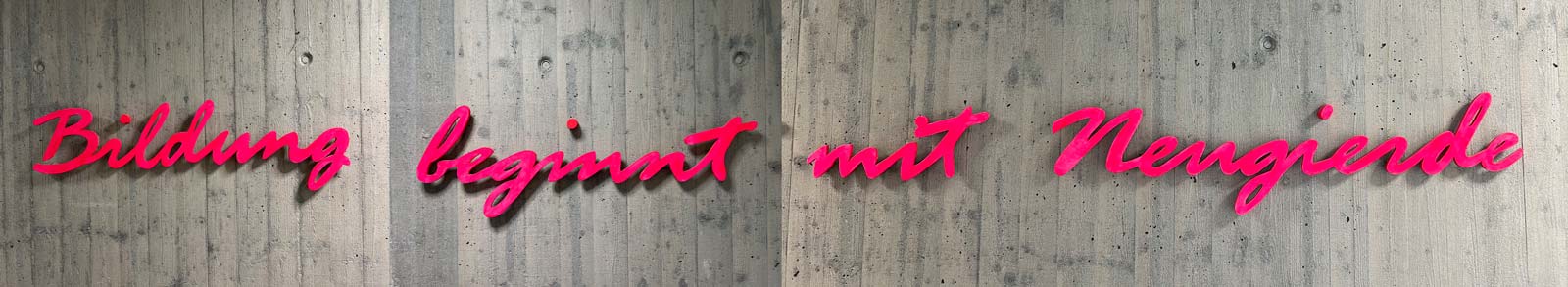 Auf einer Betonwand im Gang zwischen Lehrerzimmer und Beratungstrakt des Gymnasium Gröbenzells steht in rosa Kursivschrift „Bildung beginnt mit Neugierde“.