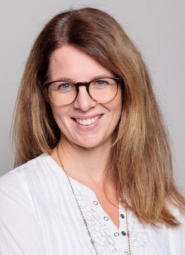 Eine Frau mit langen, braunen Haaren und Brille lächelt in die Kamera. Sie trägt eine weiße Bluse mit Knöpfen und eine Halskette und ist bereit, ihr Team zu treffen. Der Hintergrund ist schlicht und hell.